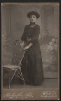 Kobieta stojąca przy krześle [fotografia]