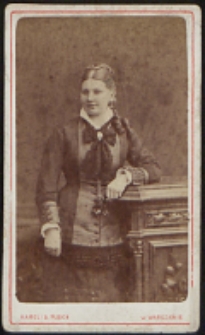 Kobieta stojąca przy komodzie [fotografia]